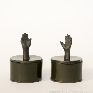 Two jars made of black clay. Each is a cylinder about 2 inches in diameter and 1.5 inches high. Each has a tiny sculpted hand and forearm protruding from the lid in place of a knob.
